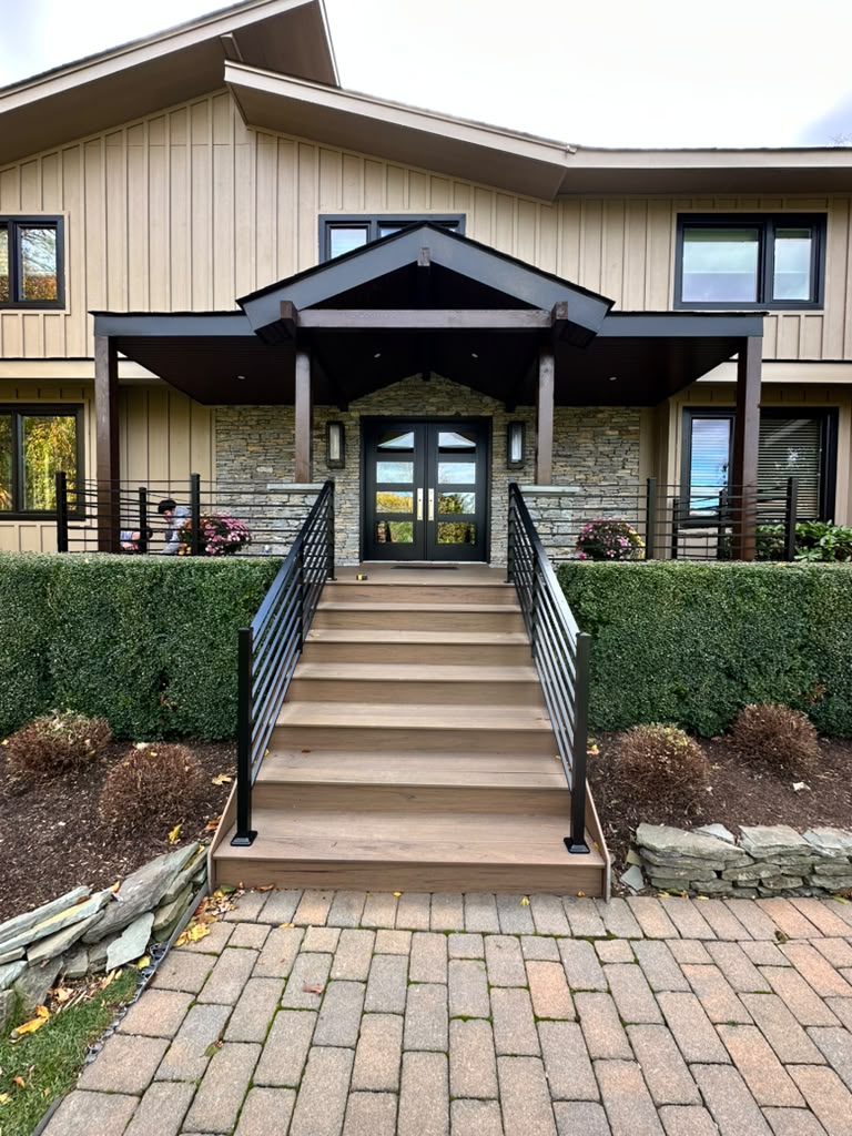 Black horizontal railing on the deck and steps of a tan board-and-batten home with paver walkway